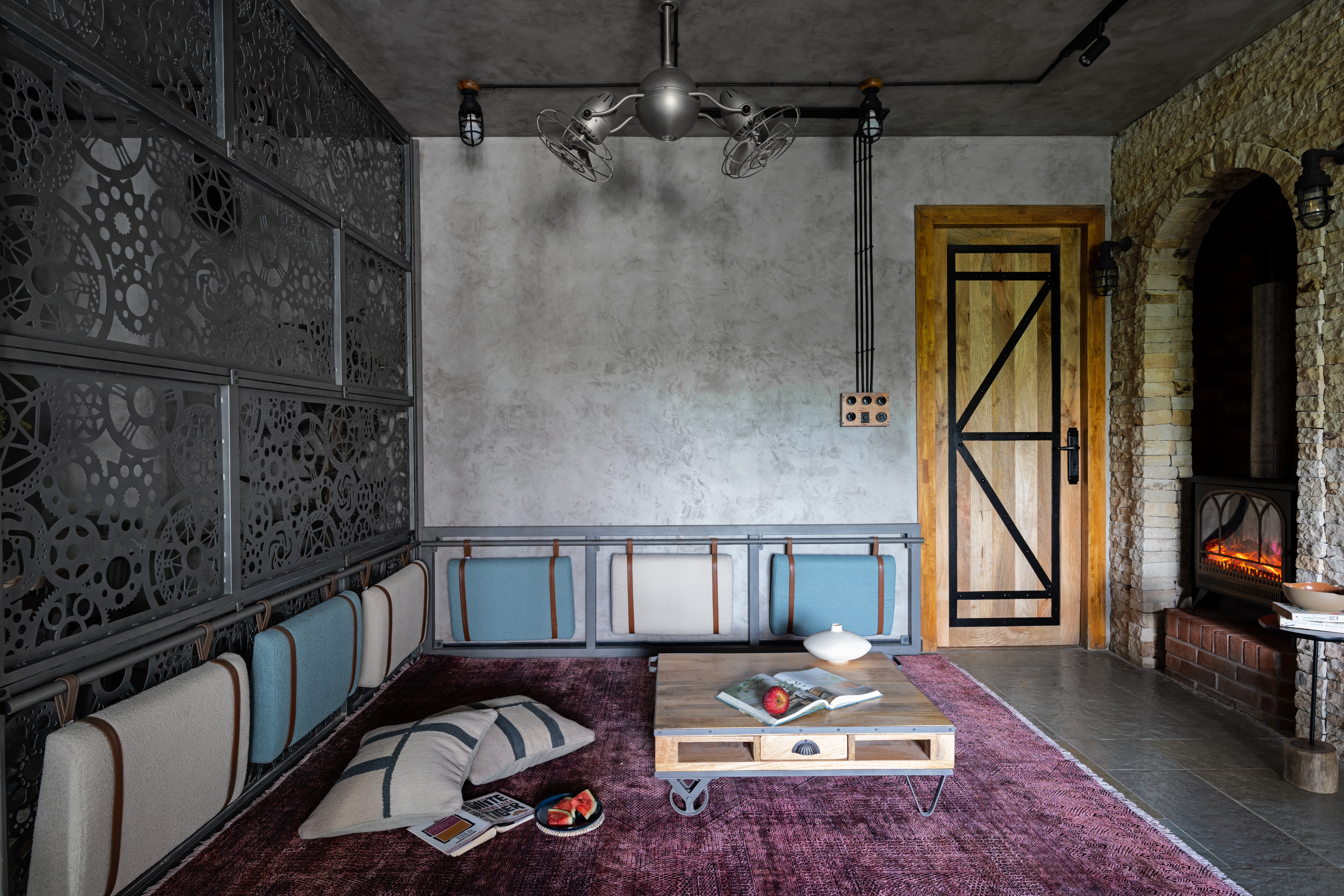 Den with a laser-cut gear-pattern steel screen wall: a low steel banquette with alternating blue and ivory cushions strapped in leather, a plum overdyed rug, a pallet-style cart table with an open book and an apple, floor cushions and a plate of watermelon, and a stove in a stone arch beyond.